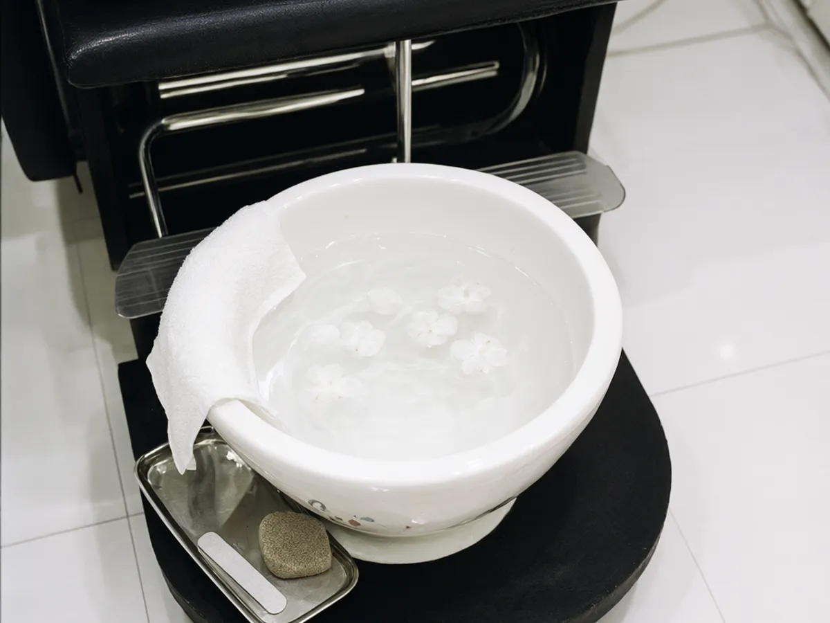 A white porcelain foot basin with floating petals on a black step in front of a pedicure chair